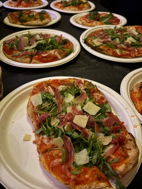 Belegte Pizzen mit Tomatensoße, Schinken, Rucola und Käse auf Papptellern.